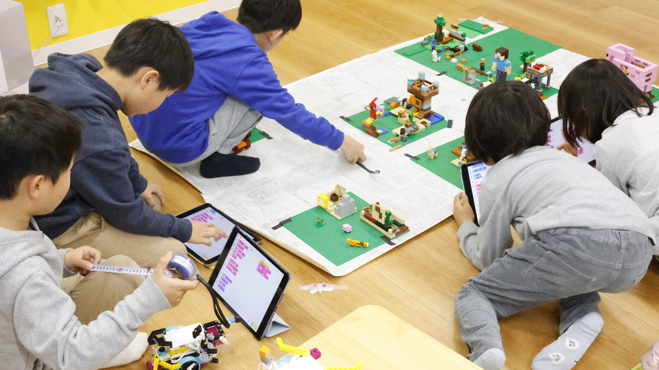 小学生たちがレゴとタブレットを使って、マインクラフトの世界をプログラミングで冒険する様子