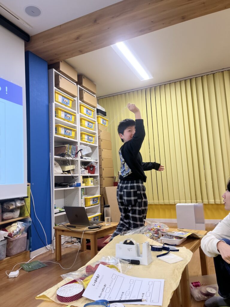 教室の中で、制作した作品の紐を高く掲げて確認している男の子