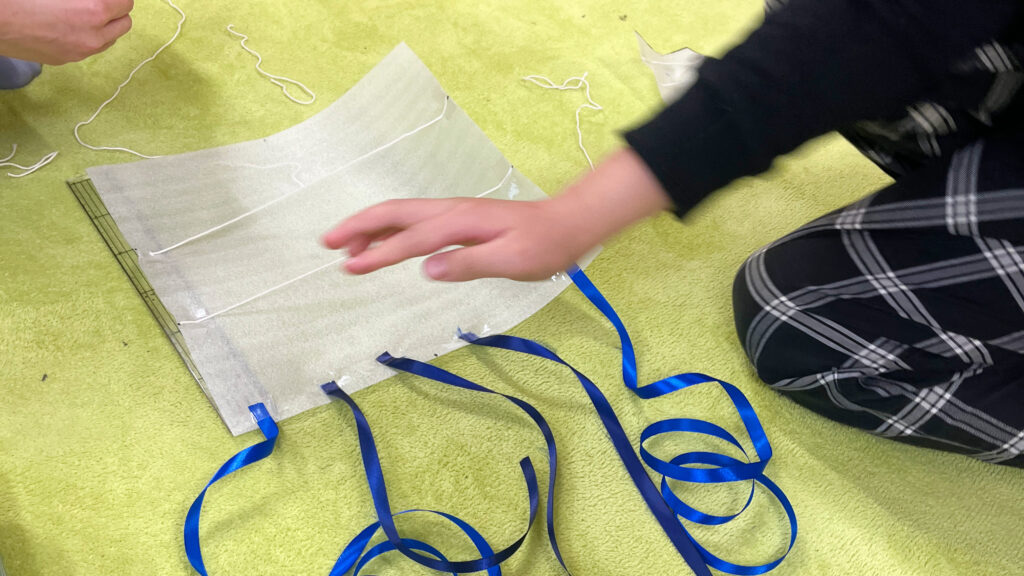 緑のマットの上で、青いリボンがついた凧（たこ）を手で押さえ、形や構造を確認している様子