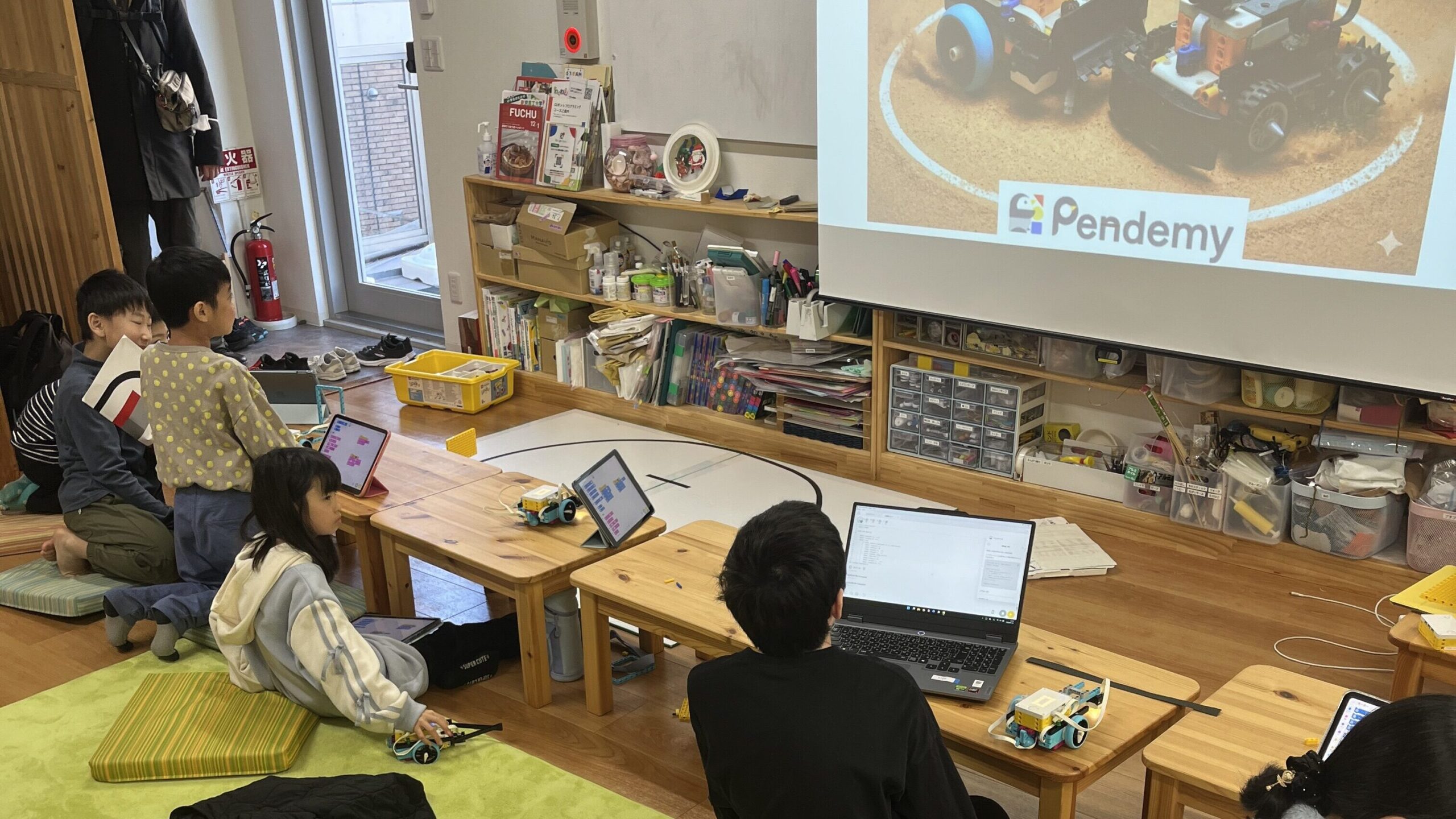 『ロボットプログラミング』と投影されたスクリーンの前で、タブレットやPCを使って課題に取り組む子供たちの様子