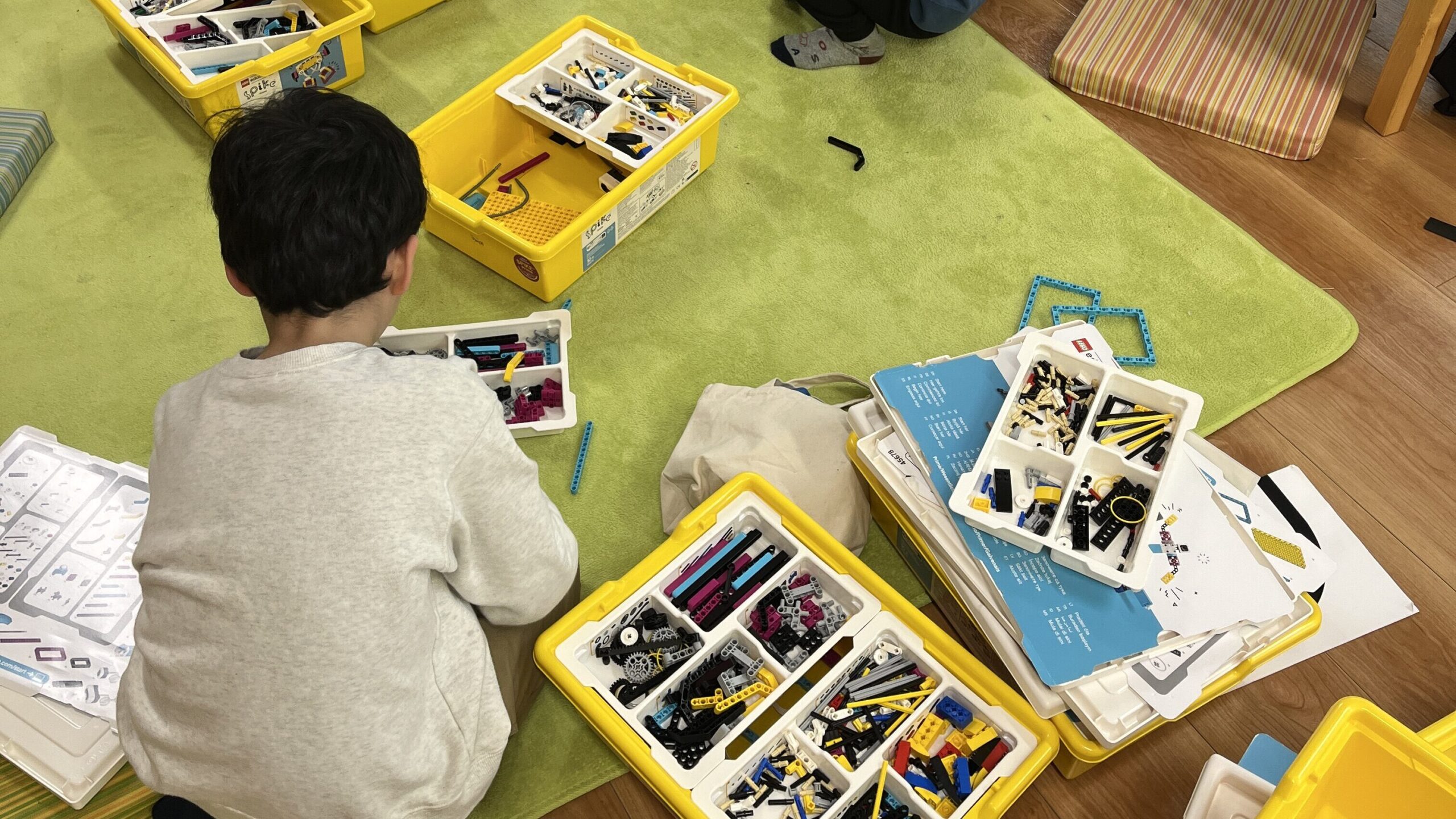 緑のラグいっぱいに黄色のレゴトレイを広げ、思い思いの体勢でロボット製作に没頭する子供たちの様子