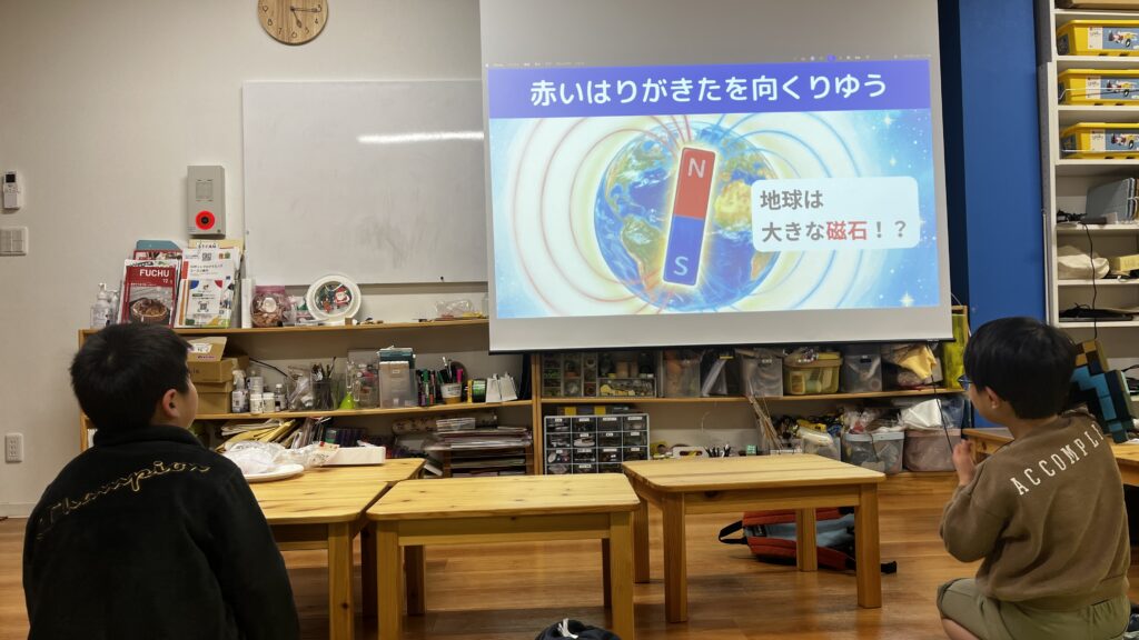 プロジェクターに映し出された「地球は大きな磁石！？」というスライドを、室内で座って見つめる2人の男の子の背中。理科の授業やワークショップの様子。