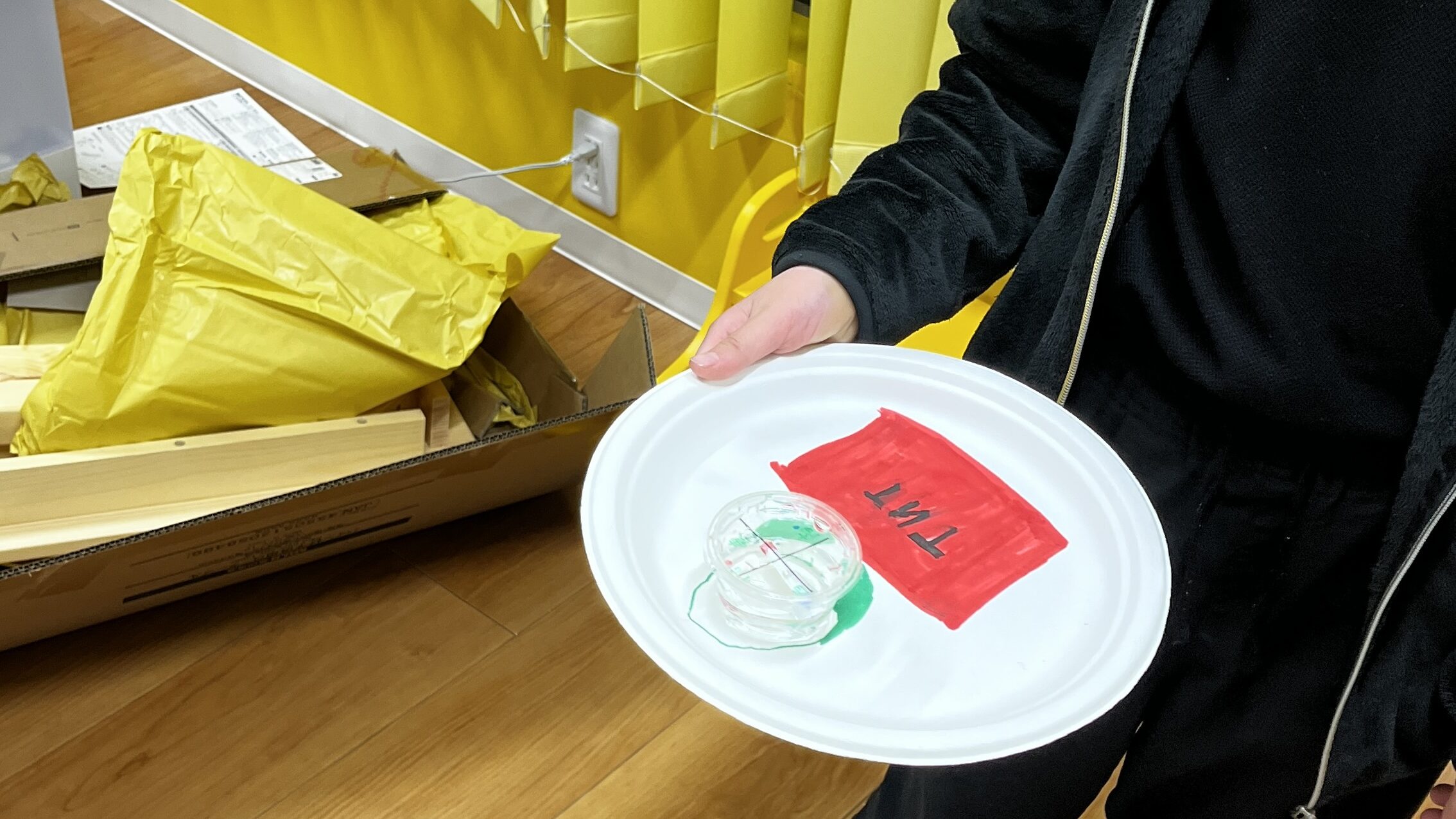 黒い服を着た男の子が、完成した手作り方位磁石と、赤い折り紙が貼られた紙皿を大切そうに両手で持っている様子。