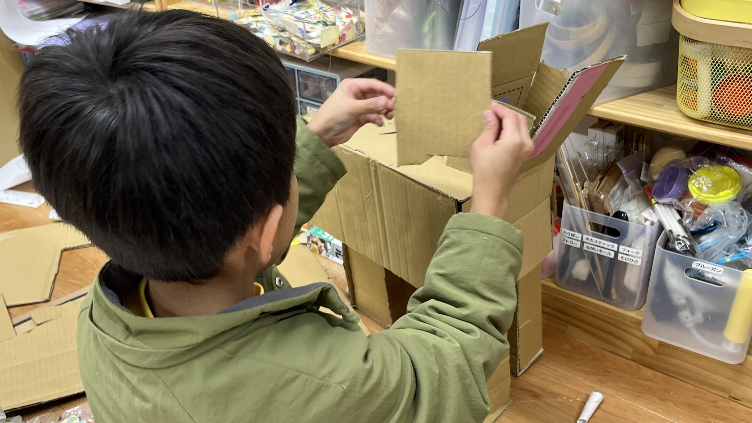 「工作資材が並ぶ棚の前で、段ボール工作に熱中する男の子の後ろ姿」