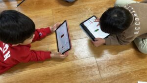 フローリングの上で、2人のお子様がそれぞれタブレットを使い、3Dモデリングソフトでペンのような物体をデザインしている様子。