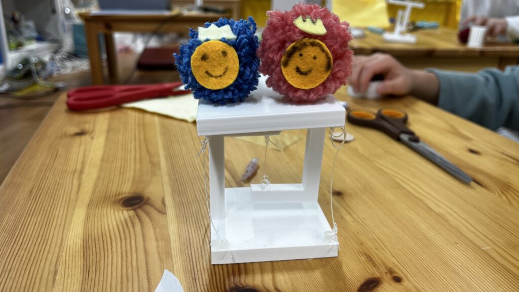 木目調のテーブルの上に置かれた、白いテンセグリティ構造の模型。その上に、青色とピンク色の毛糸で作られた2つの可愛らしいポンポン人形が乗っている。背景には工作道具や作業する人の手が写っている。