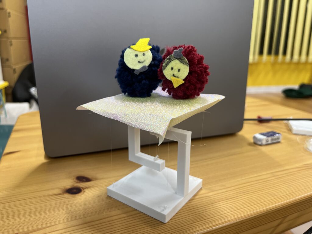 テンセグリティ構造の白い模型の上に、キラキラした金色の敷物が敷かれ、その上に毛糸のポンポンで作られたお内裏様とお雛様が並んでいる。背景にはノートパソコンがある机の上の様子が写っている。