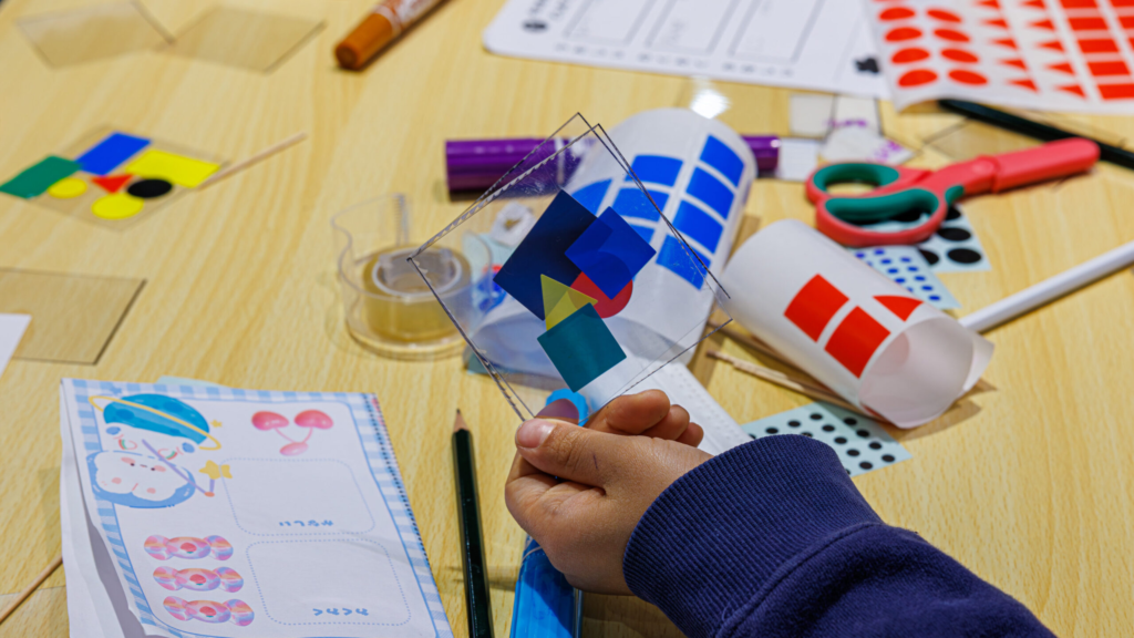 小学生が透明シートに貼った色とかたちのパーツを手に持ち、ボンボンドロップ風シールのデザインを組み立てている制作風景