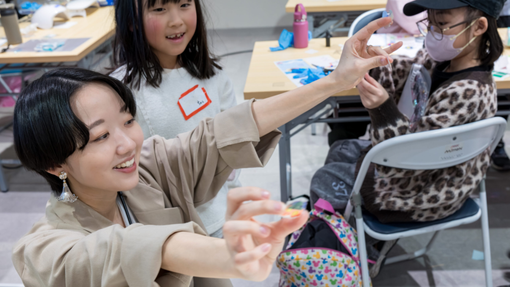 スタッフが完成したボンボンドロップ風シールを見せながら声かけし、小学生が作品を見せ合っているワークショップの様子
