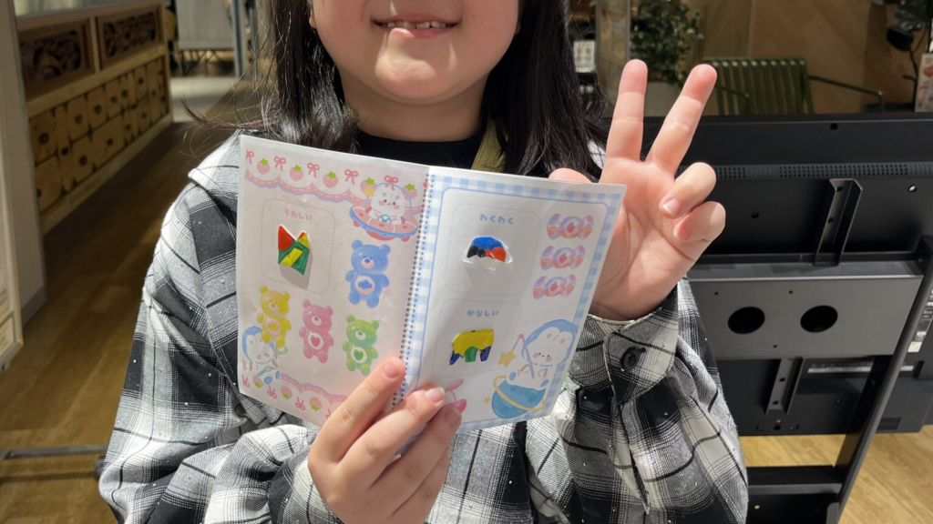 小学生が完成したボンボンドロップ風シールをシール帳に貼り、ピースサインで作品を見せている様子