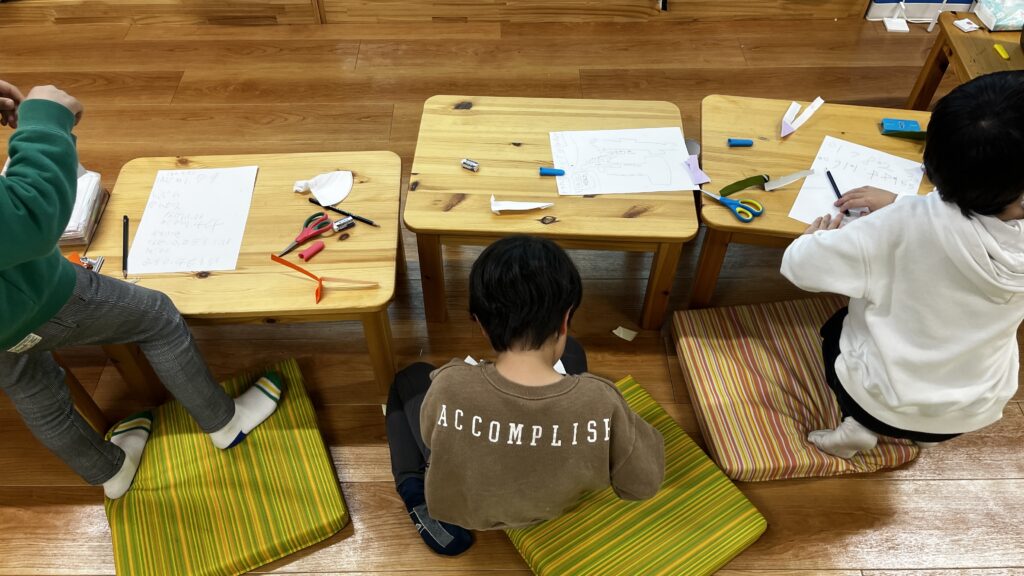 木のローテーブルに向かって、手元の紙や工作に集中する3人の子供たちの後ろ姿。テーブルには鉛筆やはさみ、実験のメモが置かれている。