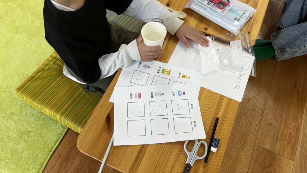 理科の実験に取り組む子供の様子。机の上には、クエン酸や塩、砂糖などのイラストが描かれたワークシート、白い粉末が入った袋、コップ、文房具が置かれています。