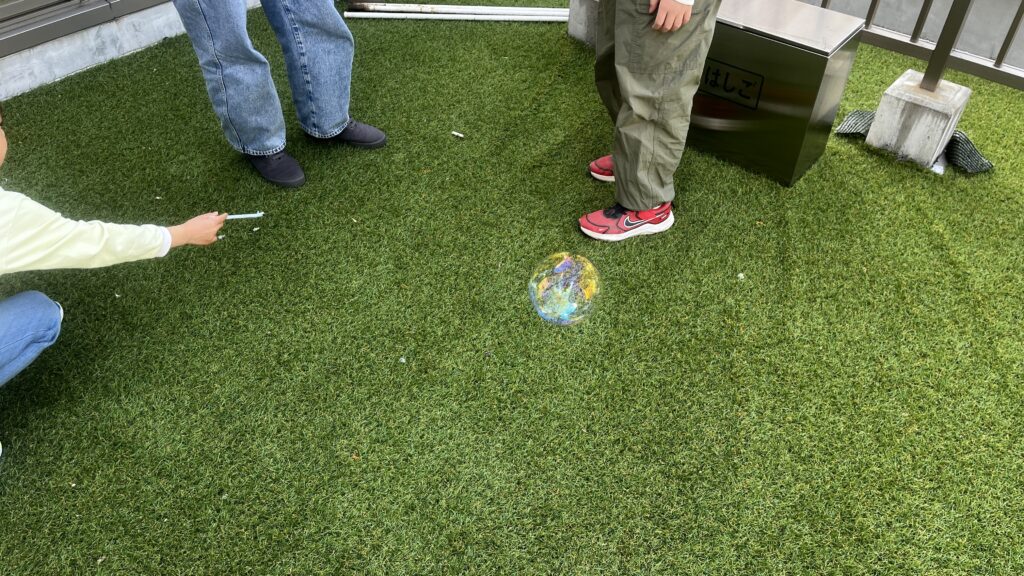 人工芝の上で浮かんでいる大きなシャボン玉と、それを見守る子供たちの足元