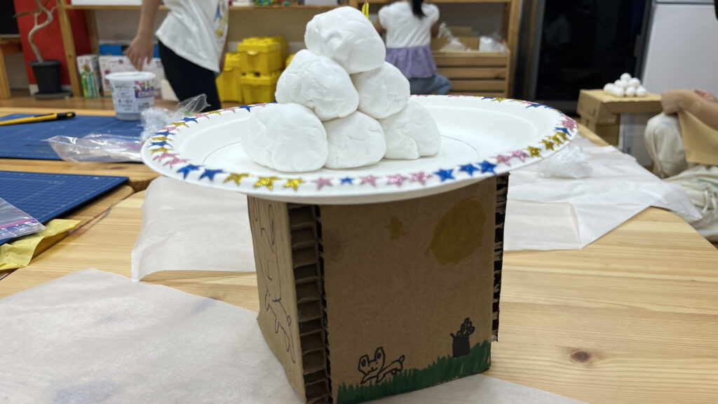 星のシールで飾った紙皿と段ボールの台で作った三方に、紙粘土の月見だんごを積んだ小学生の工作