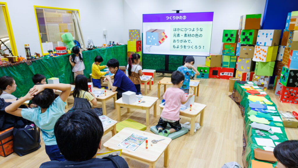 :「つくりかた③」のスライドを前に、低いテーブルで色紙を貼りながらブロックを作る子どもたちと見守る保護者