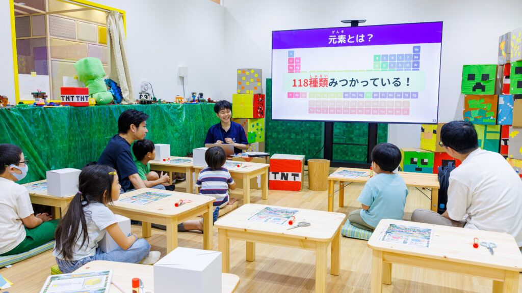「元素とは? 118種類みつかっている!」のスライドを前に、スタッフの話を聞く小学生と保護者