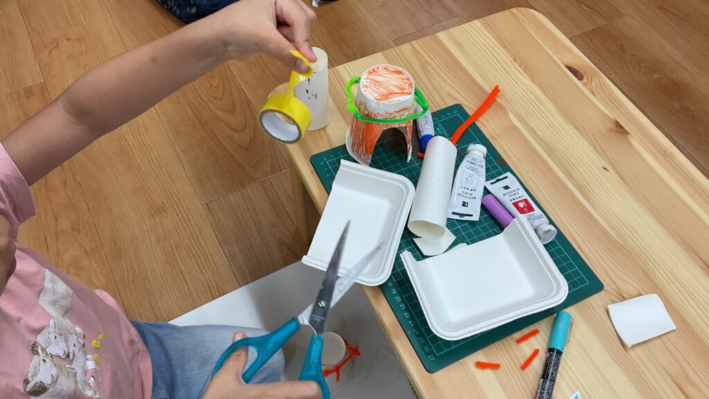 年長の女児が、生鮮品用包装パックや紙コップなどを用いて工作をしている工作教室の様子。