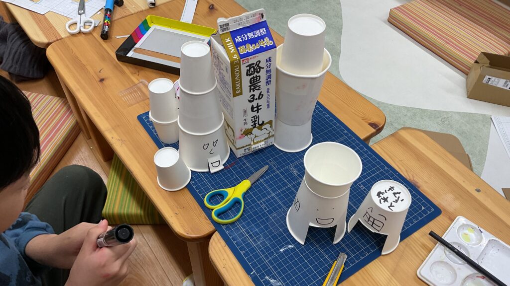 牛乳パックや紙コップを組み合わせたりして顔が描かれた家を作る小学生男児。