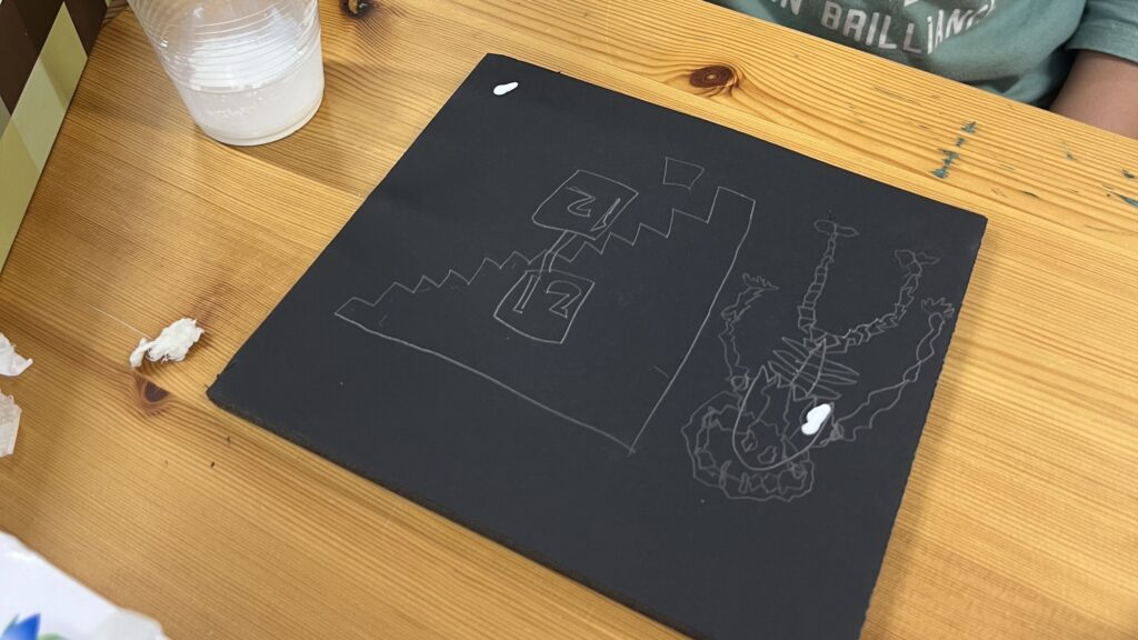 小学生向け実験教室で、黒い台紙に描いたお城やドラゴンの下絵に、割り箸を使って白い材料を塗り、光る立体アートを制作する様子