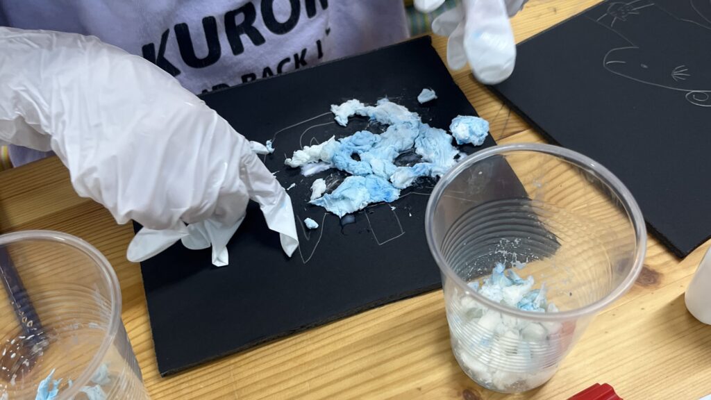 小学生が手袋を着用し、水色と白の材料を黒い台紙に貼り付けて立体アートを制作する実験教室の様子